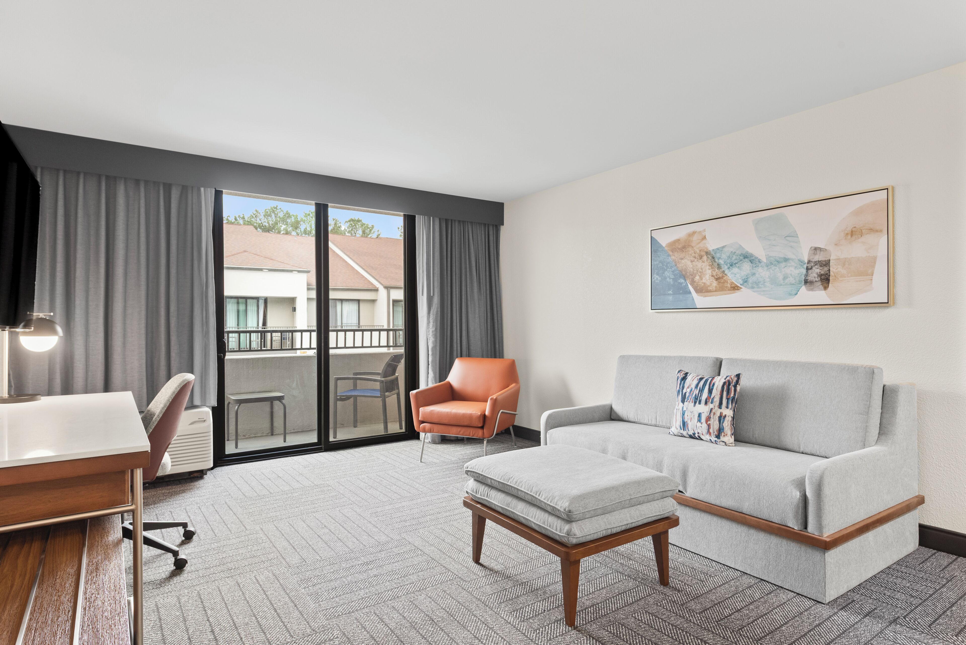 A modern hotel room with a grey sofa, a small table, a chair, and a large window with curtains.
