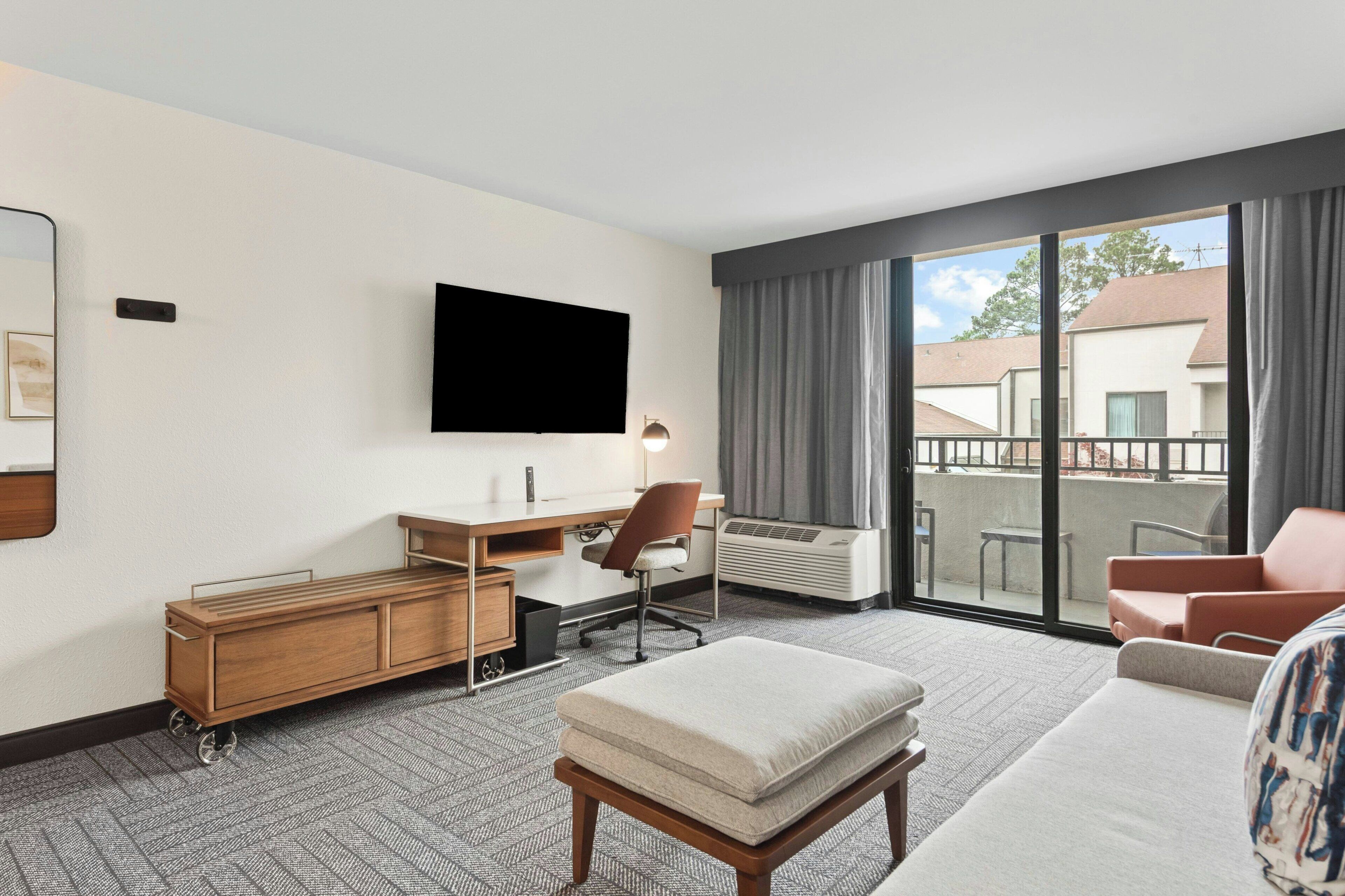 A hotel room with a flat-screen TV, a desk, a chair, a sofa, and a window with a view of buildings.