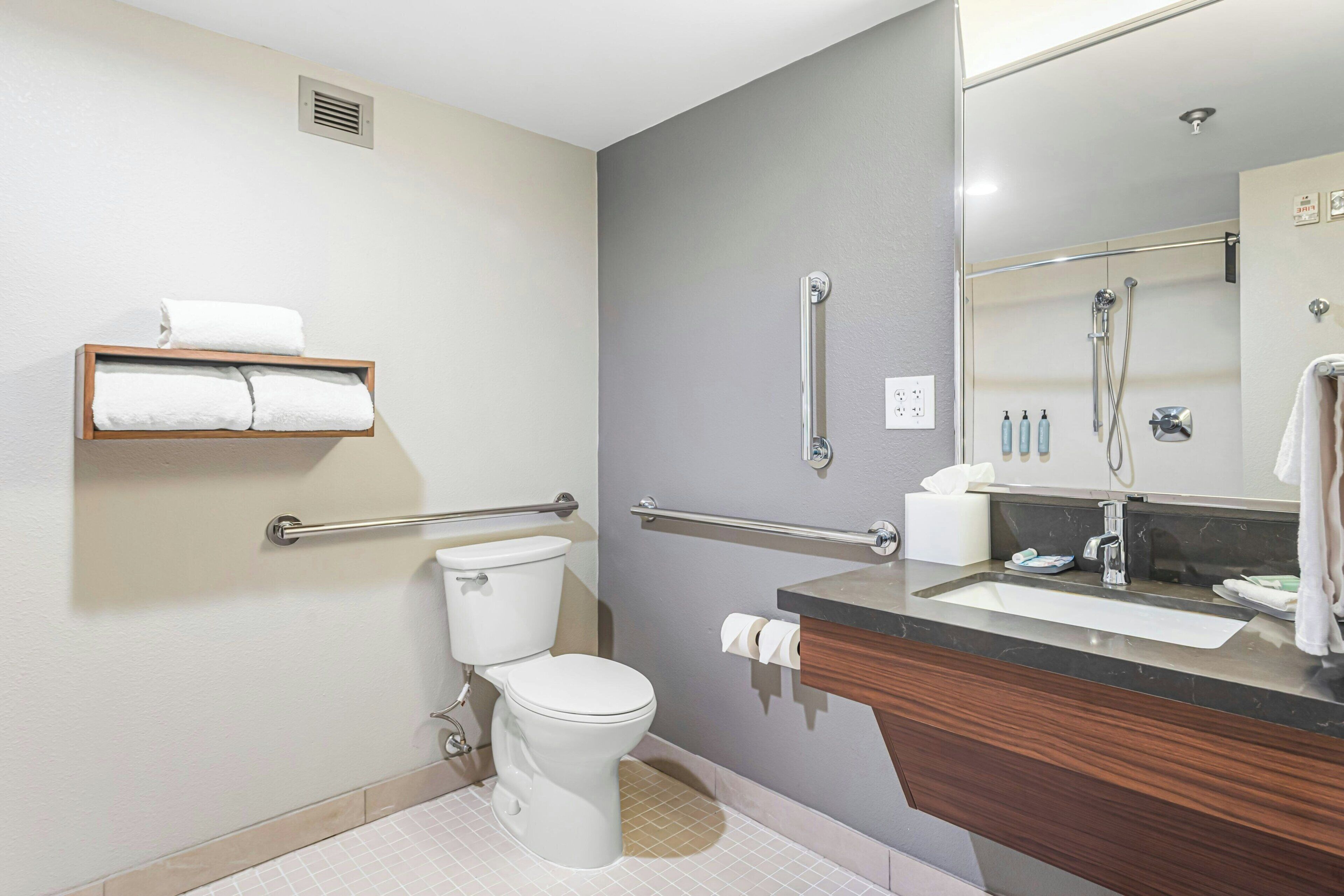 A modern bathroom with a toilet, grab bars, a towel rack, a sink with a mirror, and a shower area.