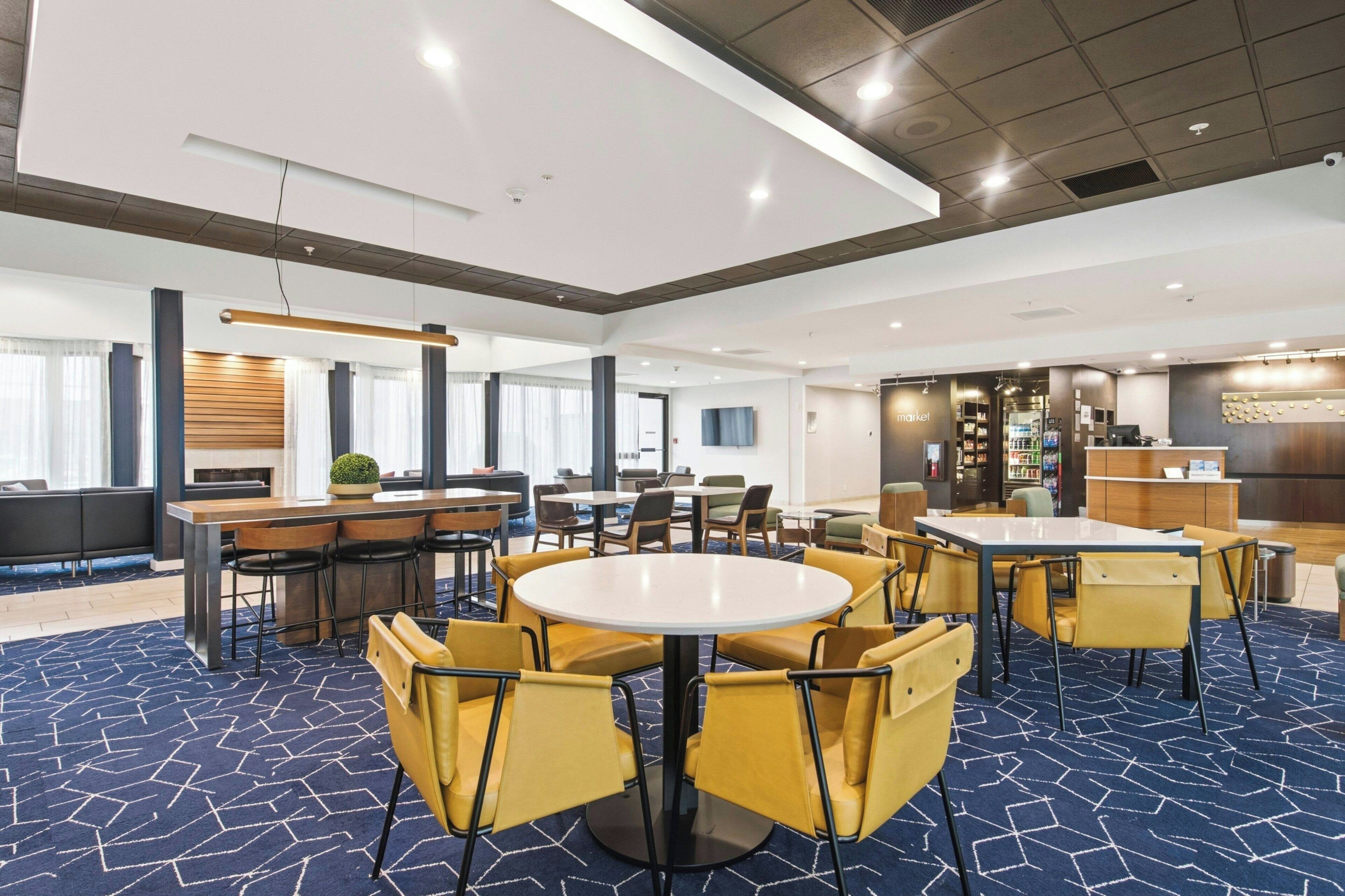 A spacious hotel lobby with multiple seating areas, including round tables and chairs, a reception desk, and a bar area.