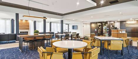A spacious hotel lobby with multiple seating areas, including round tables and chairs, a reception desk, and a bar area.