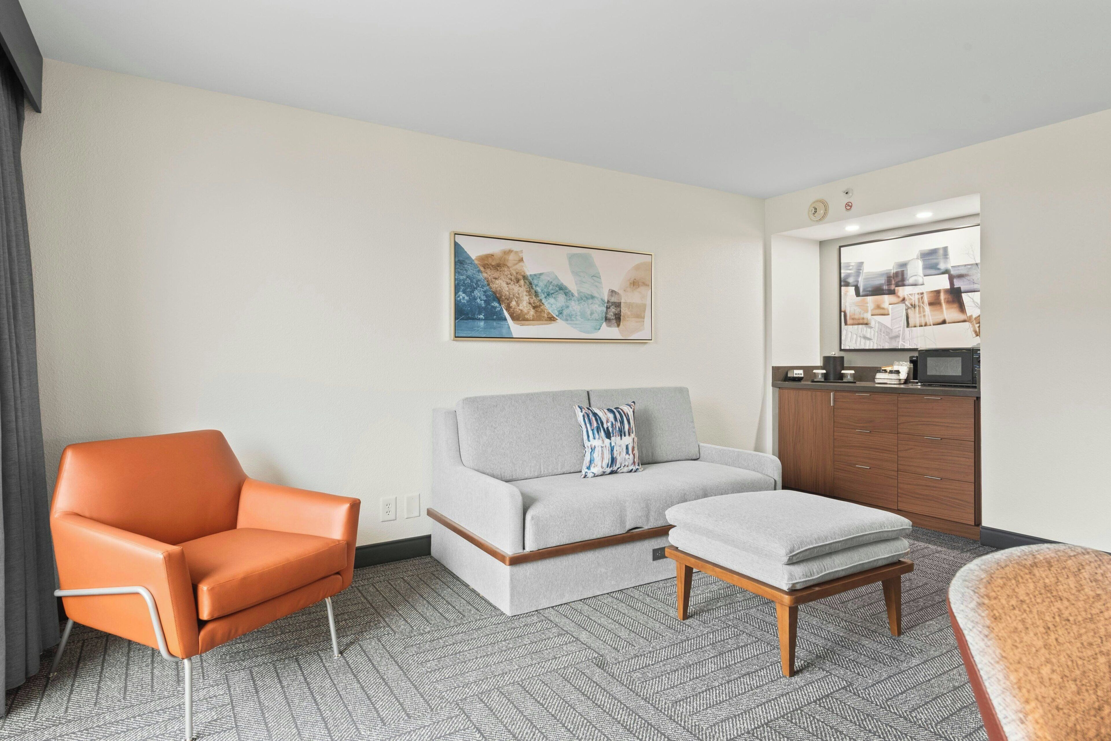 A hotel room with a sofa, an ottoman, a chair, a dresser, and a painting on the wall.