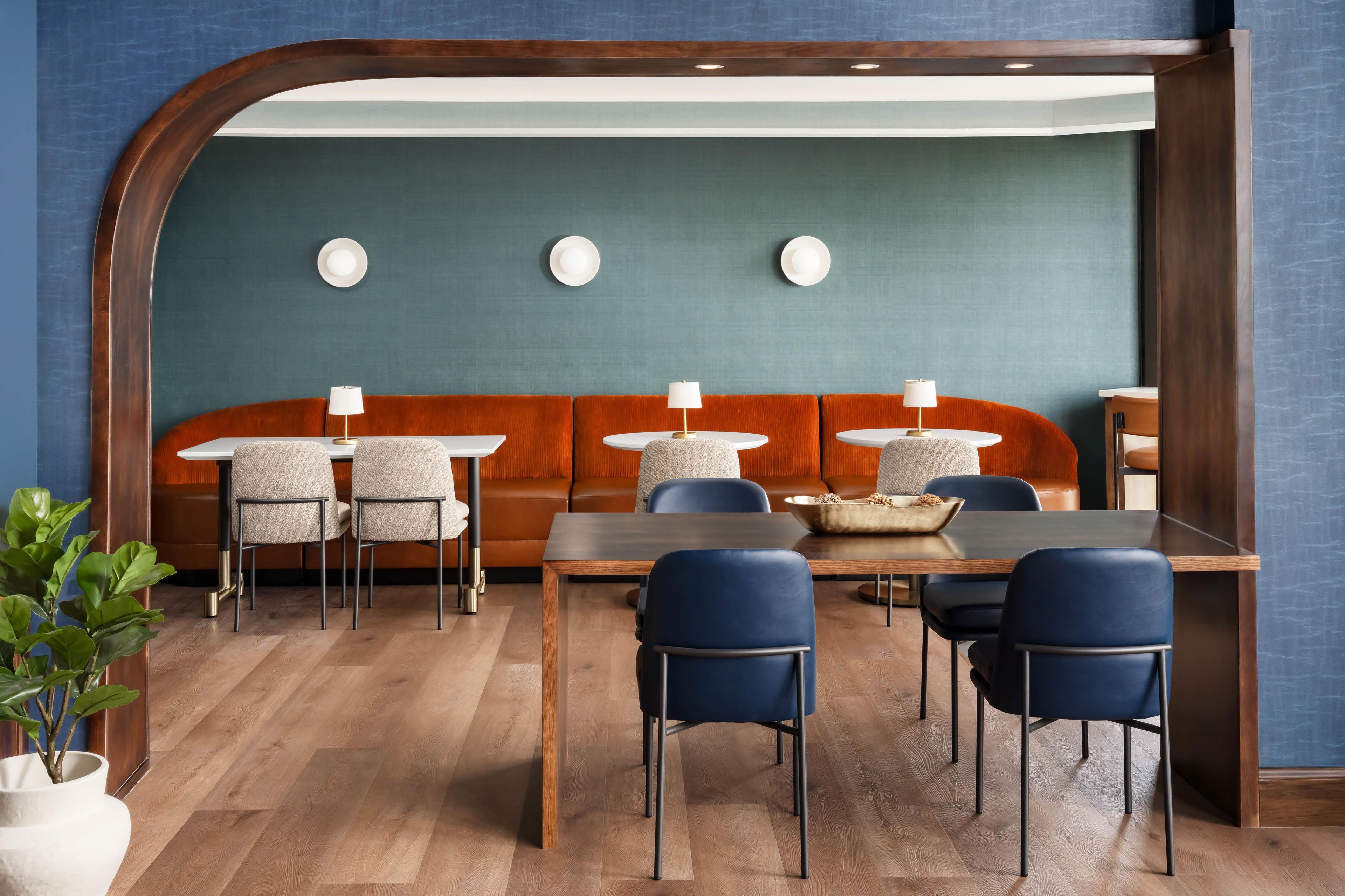 A modern interior with a wooden dining table, blue chairs, a sofa, and a wall-mounted light fixture.