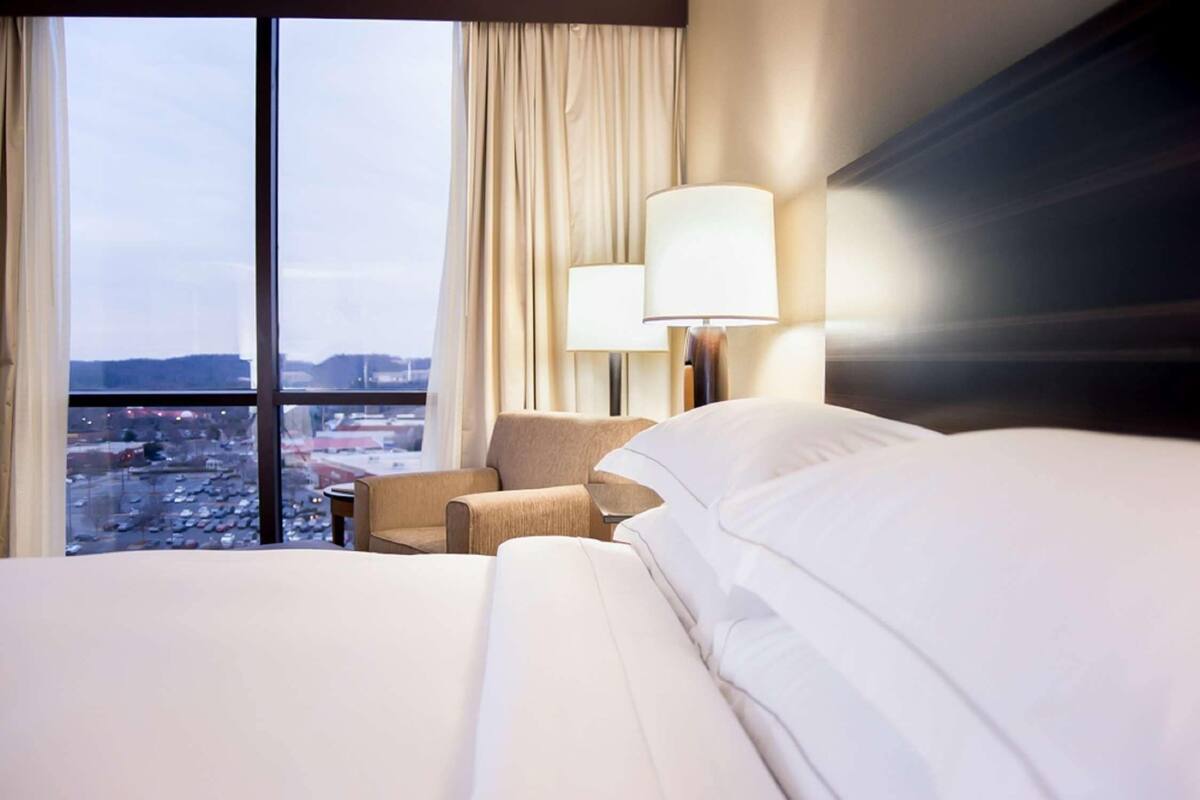 A hotel room with a large bed, a chair, a lamp, and a view of the city through large windows.