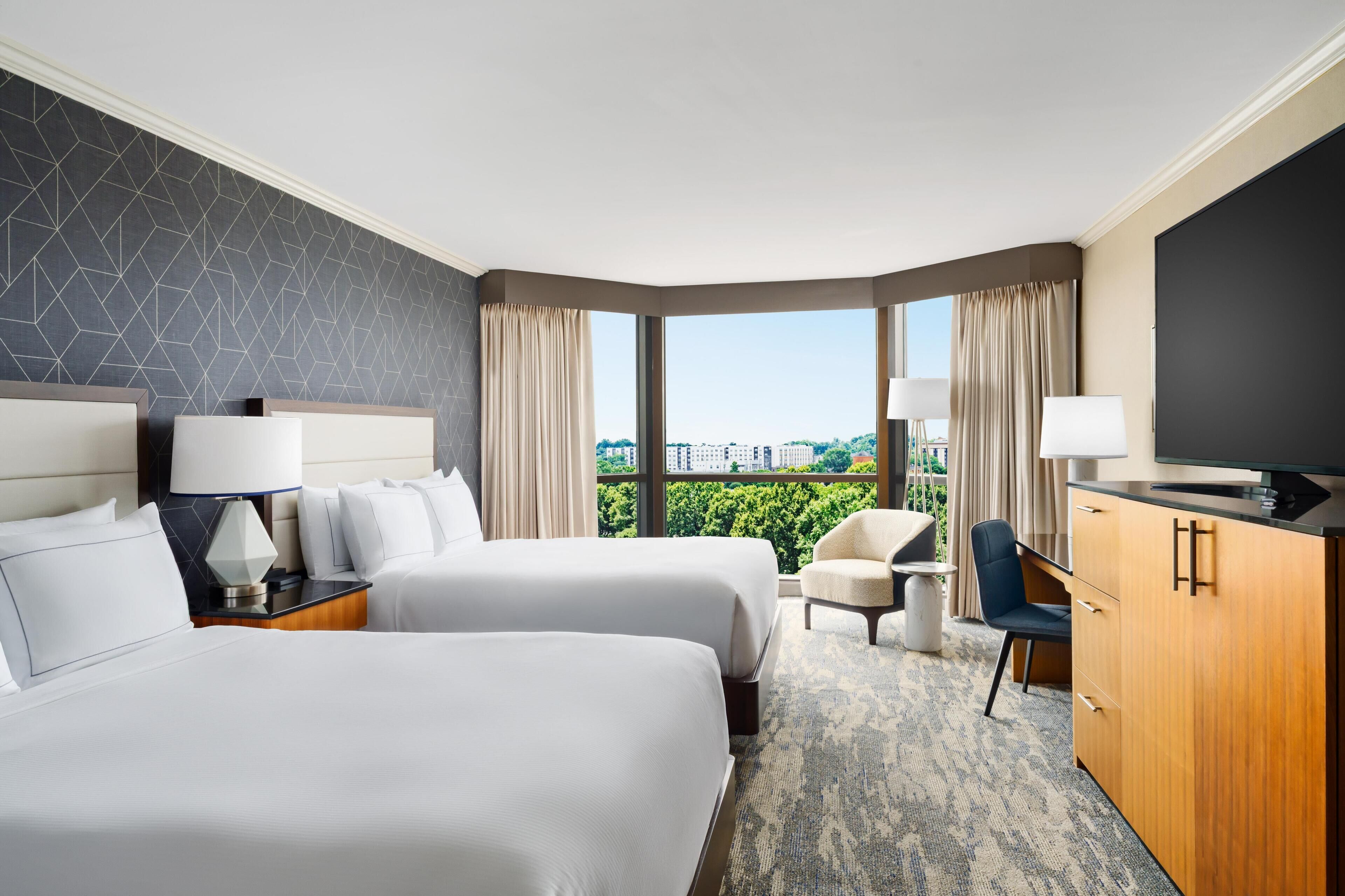 A hotel room with two beds, a desk, a chair, a TV, and a large window with a city view.
