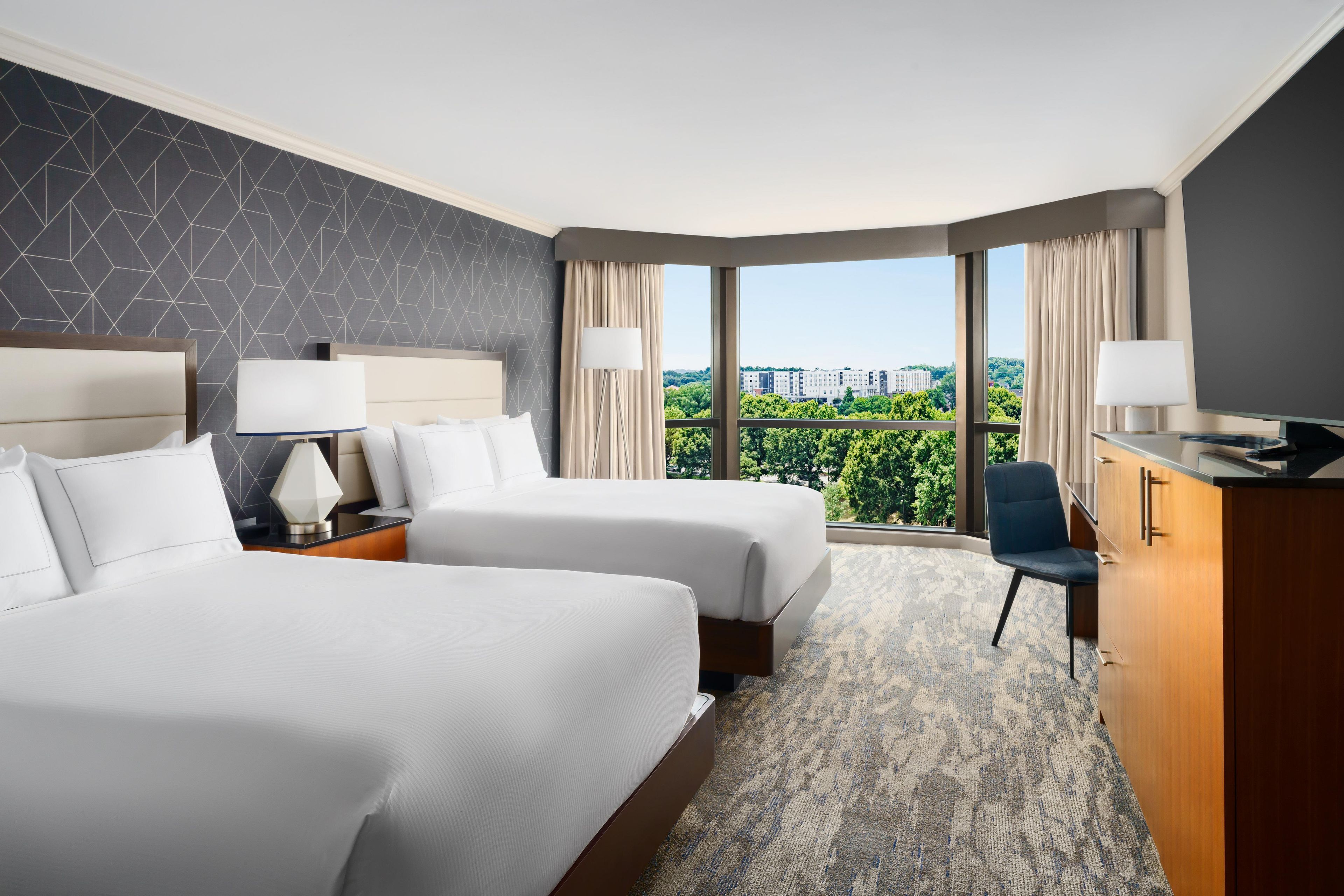 Ein Hotelzimmer mit zwei Betten, einem Schreibtisch, einem Sessel und einem großen Fenster mit Blick auf die Stadt.