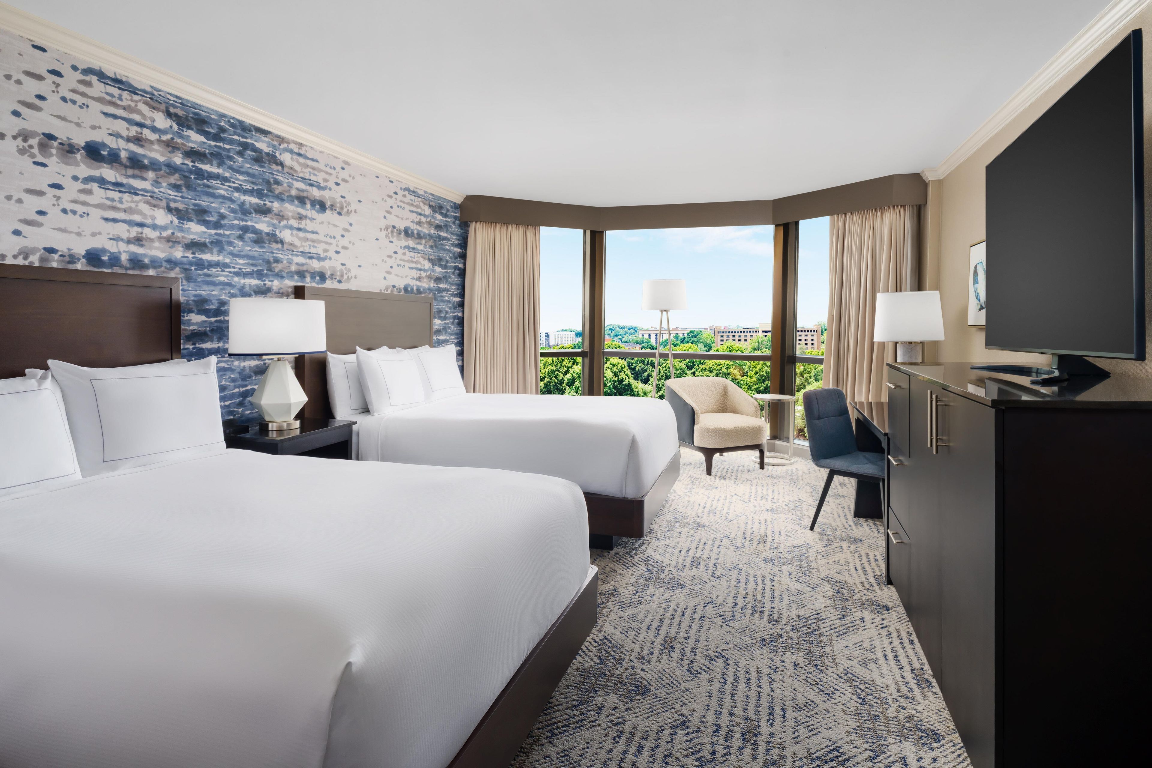 A hotel room with two beds, a desk, a chair, a TV, and a large window with a view of buildings and greenery.