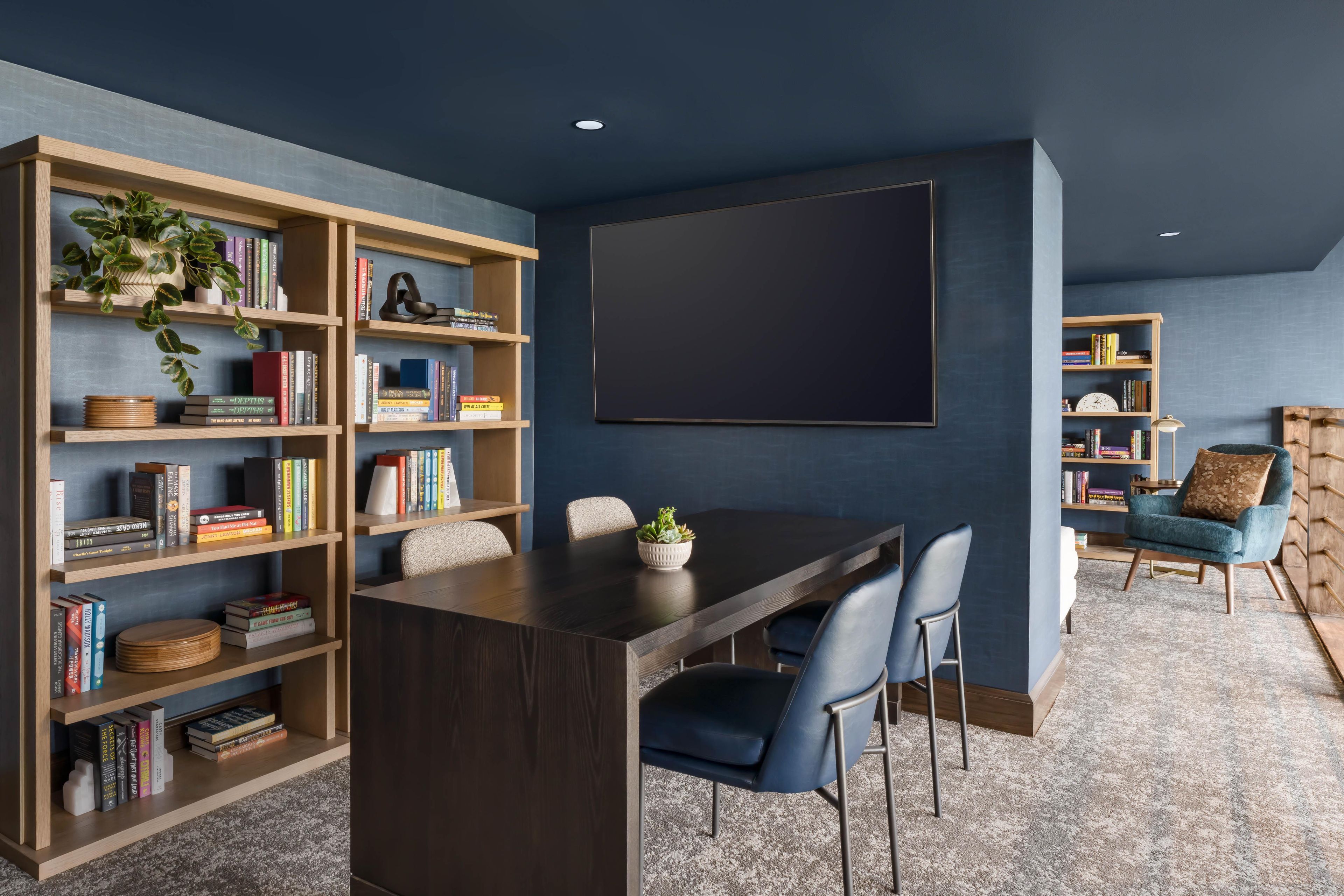 A modern living room with a dark wooden dining table, blue chairs, and a large bookshelf filled with books and decorative items.