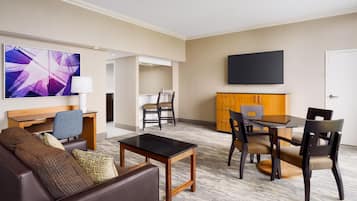 A hotel room with a living area, a dining table, a sofa, a flat-screen TV, and a minibar.