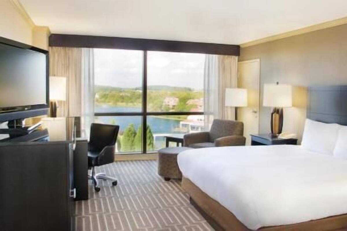 A hotel room with a large bed, a desk with a TV, a chair, a lamp, and a view of a lake.