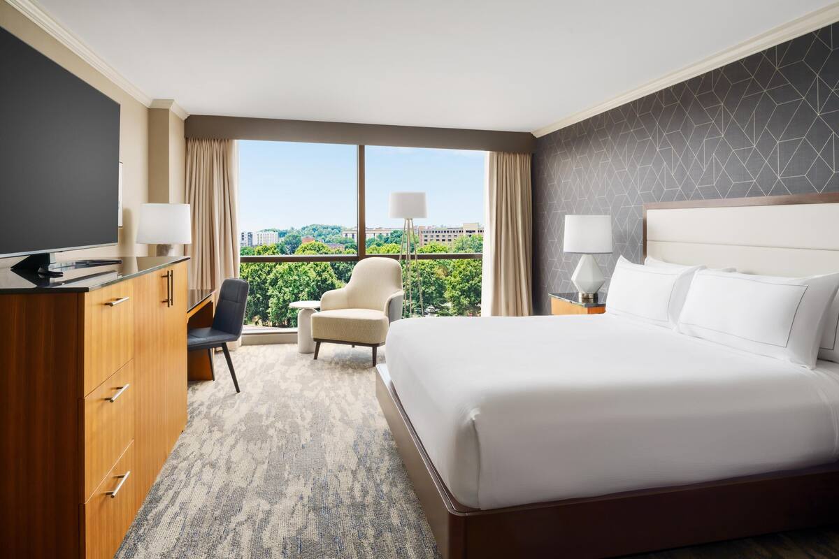 A hotel room with a bed, desk, chair, and a view of greenery and buildings.