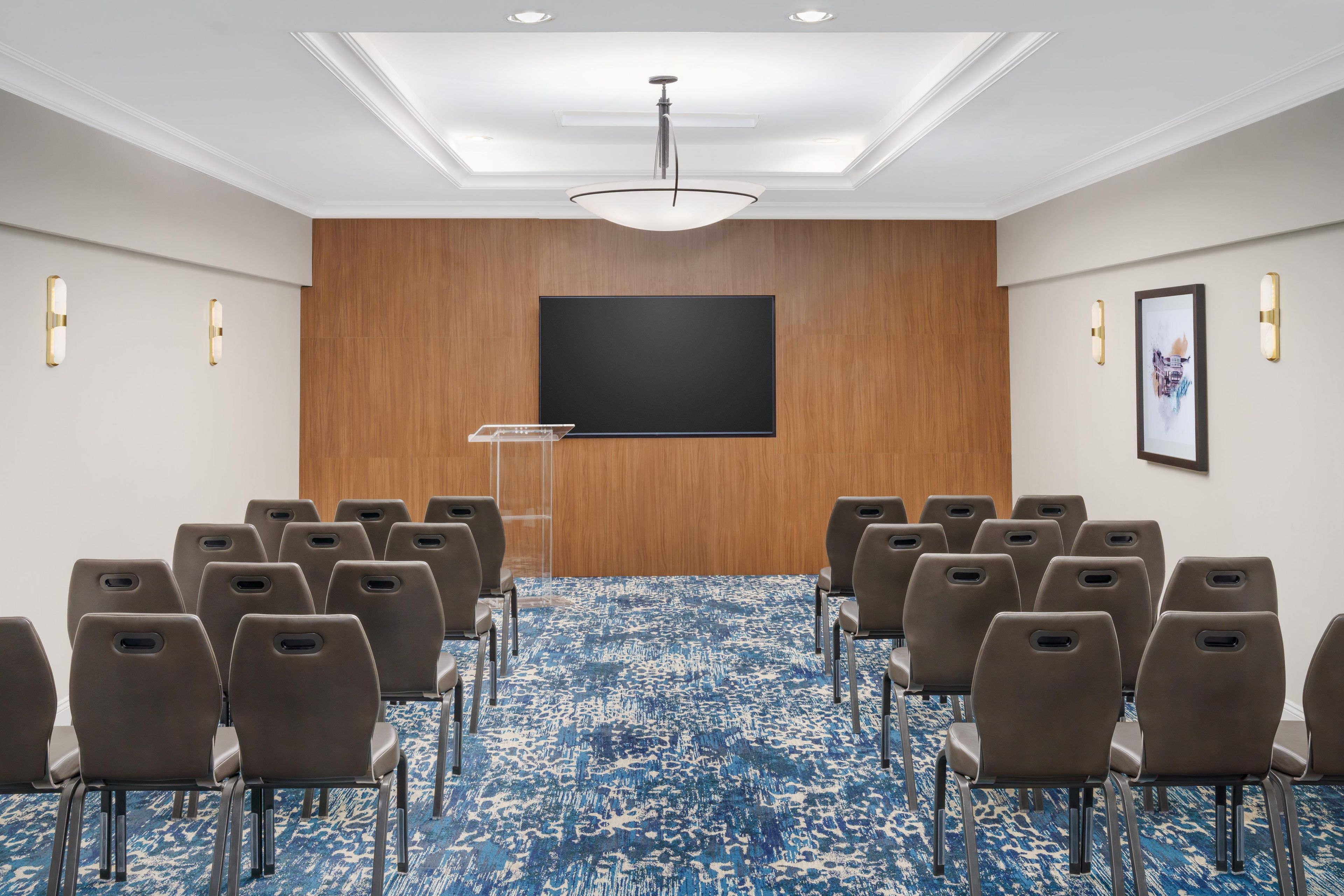 A conference room with rows of chairs, a large flat-screen TV, a podium, and a patterned carpet.