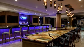 A bar area with a long counter, multiple bar stools, and hanging pendant lights. - Hilton Charlotte University Place (Charlotte)