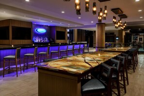 A bar area with a long counter, multiple bar stools, and hanging pendant lights. - Hilton Charlotte University Place (Charlotte)