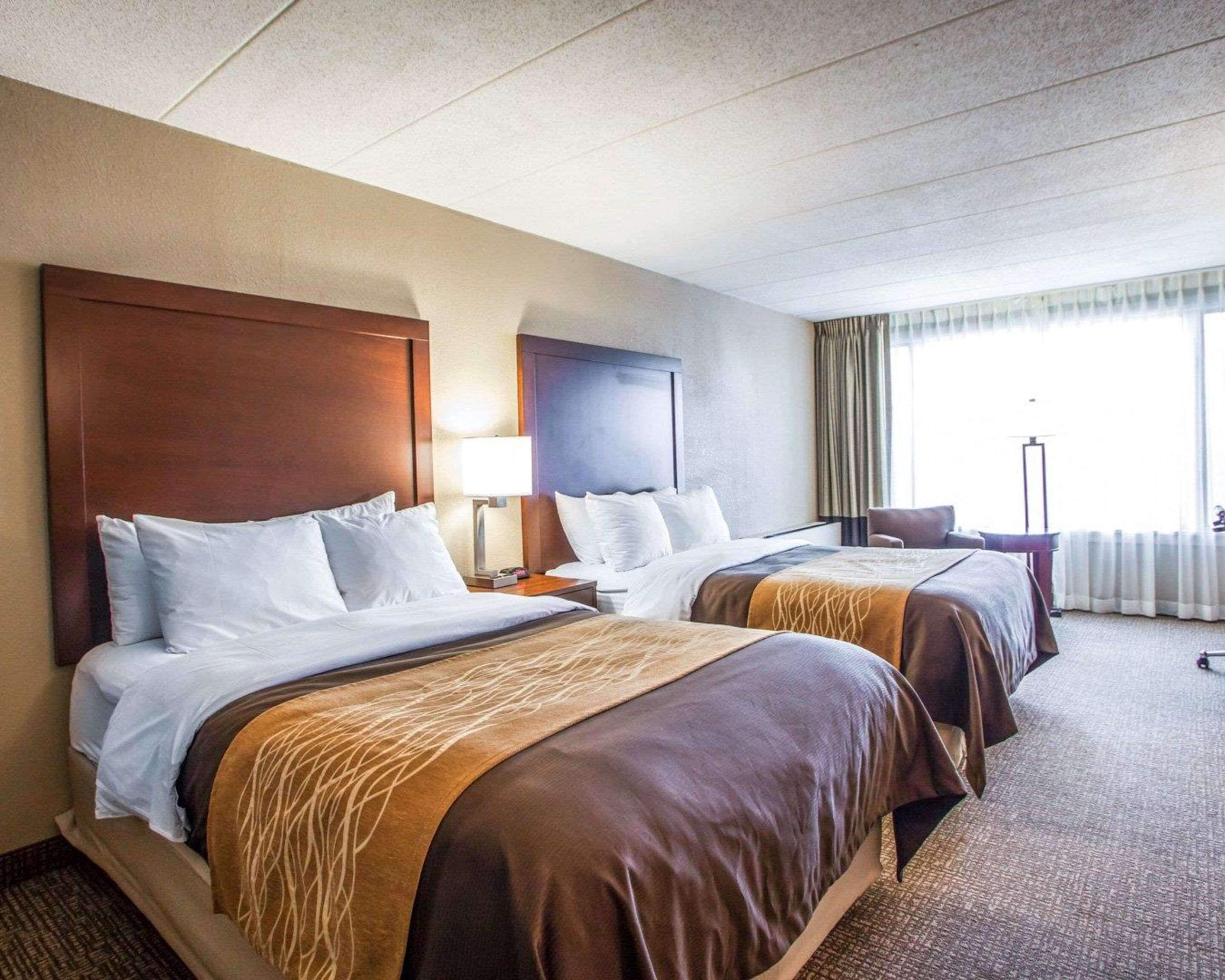 A hotel room with two beds, a nightstand with a lamp, a chair, and a large window with curtains.