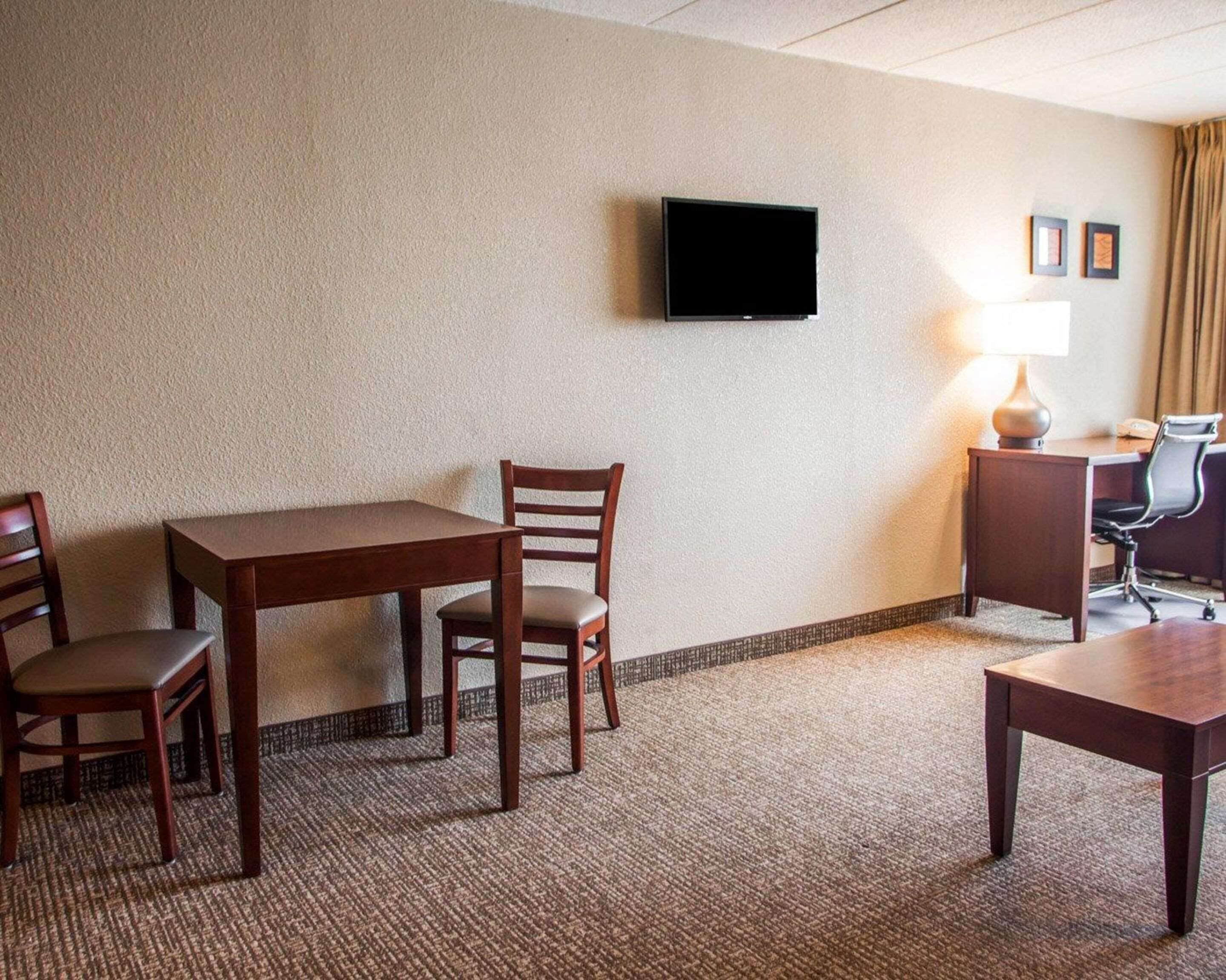 A hotel room with a wooden table, two chairs, a TV mounted on the wall, a desk with a lamp, and a small rug.