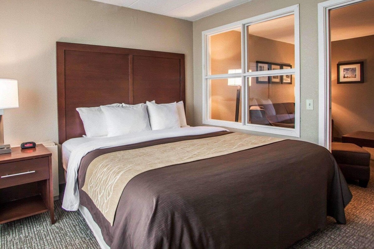 Una habitación de hotel con una cama grande, cabecera de madera, una mesita de noche con lámpara, ventana con vistas y una puerta que da a otra habitación.