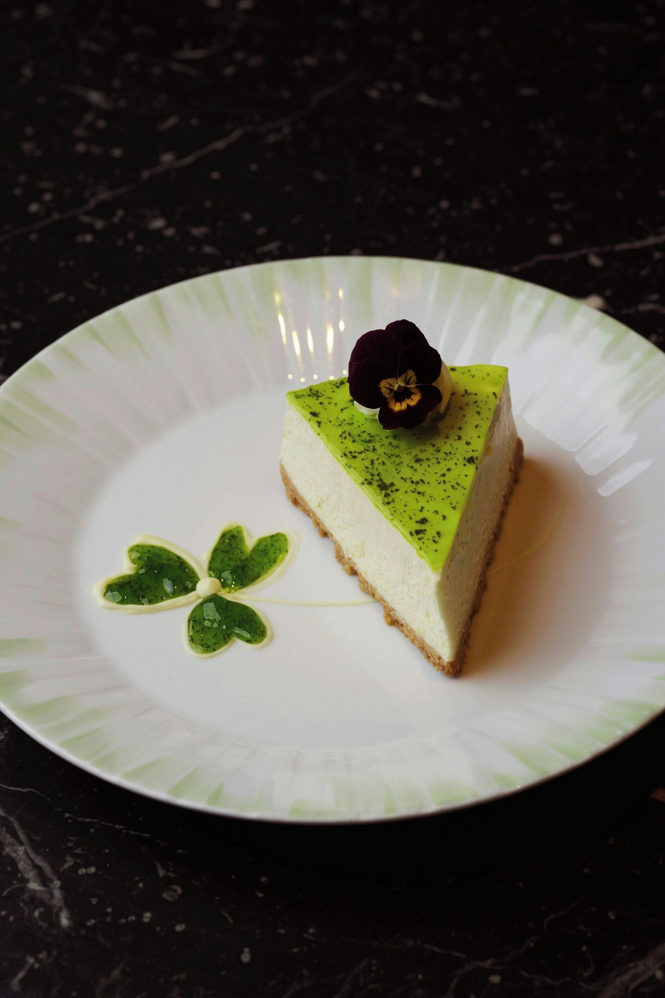 Ein Stück Käsekuchen auf einem weißen Teller, mit einer Zierblume darauf.