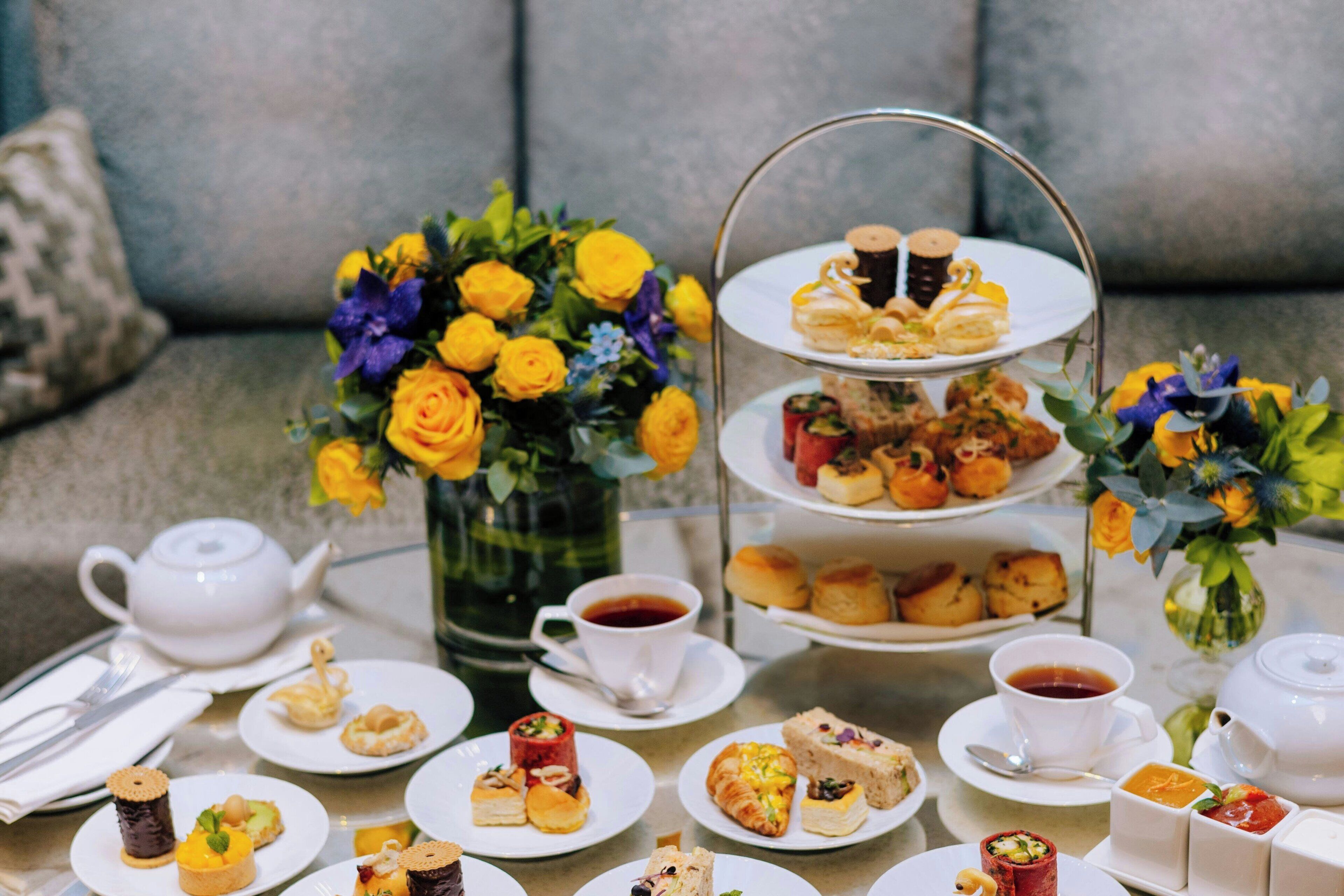 A tea service with multiple tiers of pastries and sandwiches on a table, accompanied by a vase of yellow roses and a teapot.