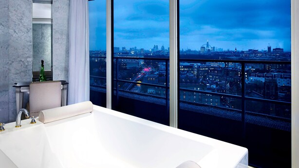 A modern bathroom with a large bathtub, a mirror, and a view of a cityscape through large windows. - The Park Tower Knightsbridge, A Luxury Collection Hotel (London)