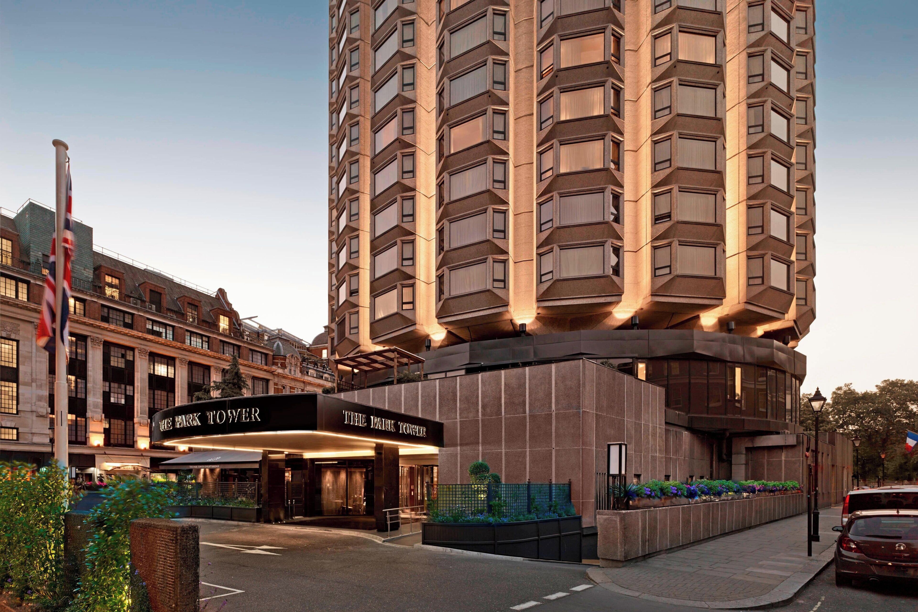 A high-rise hotel with a modern design, featuring a prominent entrance with a sign, a flag, and a well-maintained garden area.