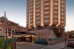 A high-rise hotel with a modern design, featuring a prominent entrance with a sign, a flag, and a well-maintained garden area. - The Park Tower Knightsbridge, A Luxury Collection Hotel (London)