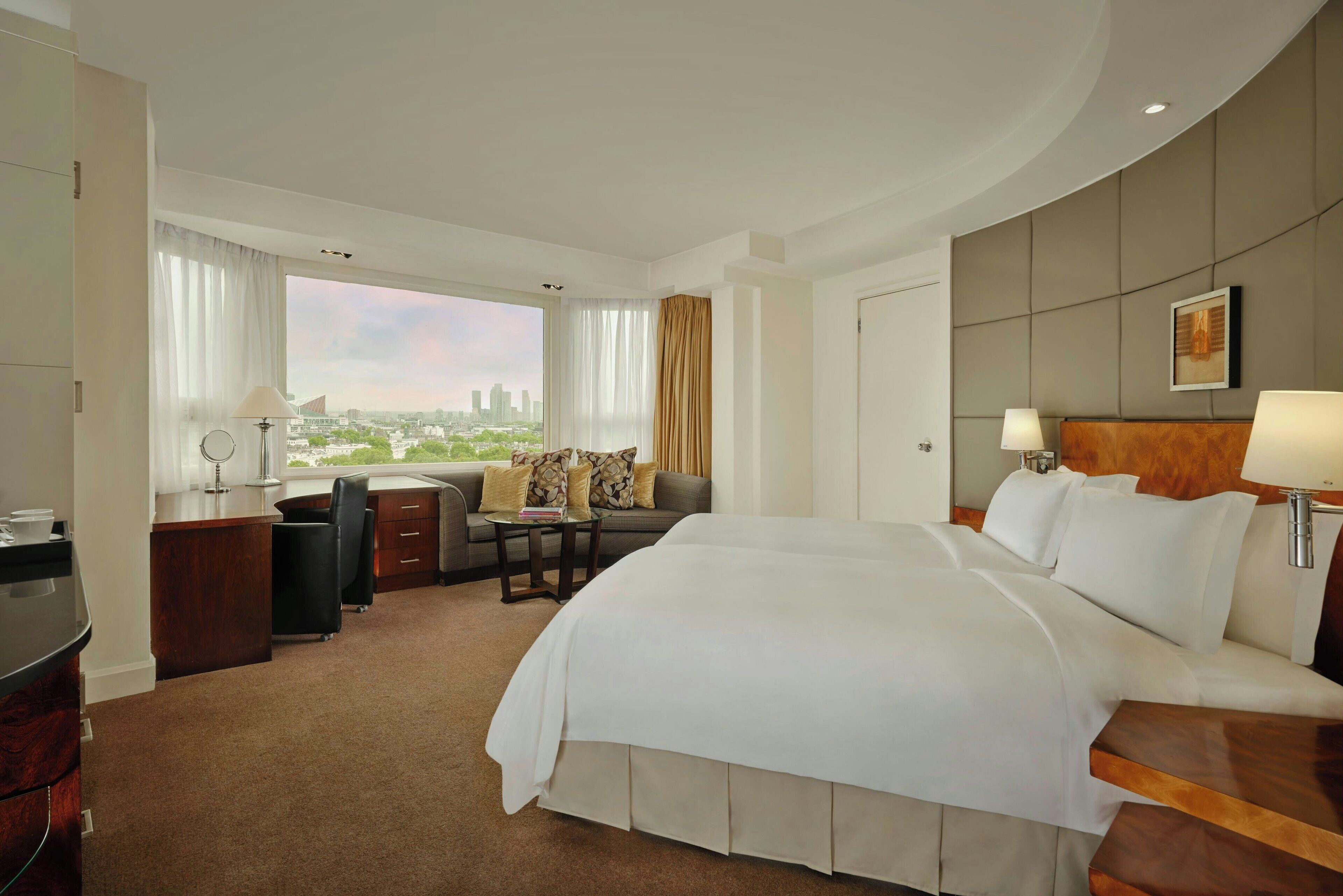 A hotel room with a large bed, a desk with a chair, a sofa, and a view of the cityscape.