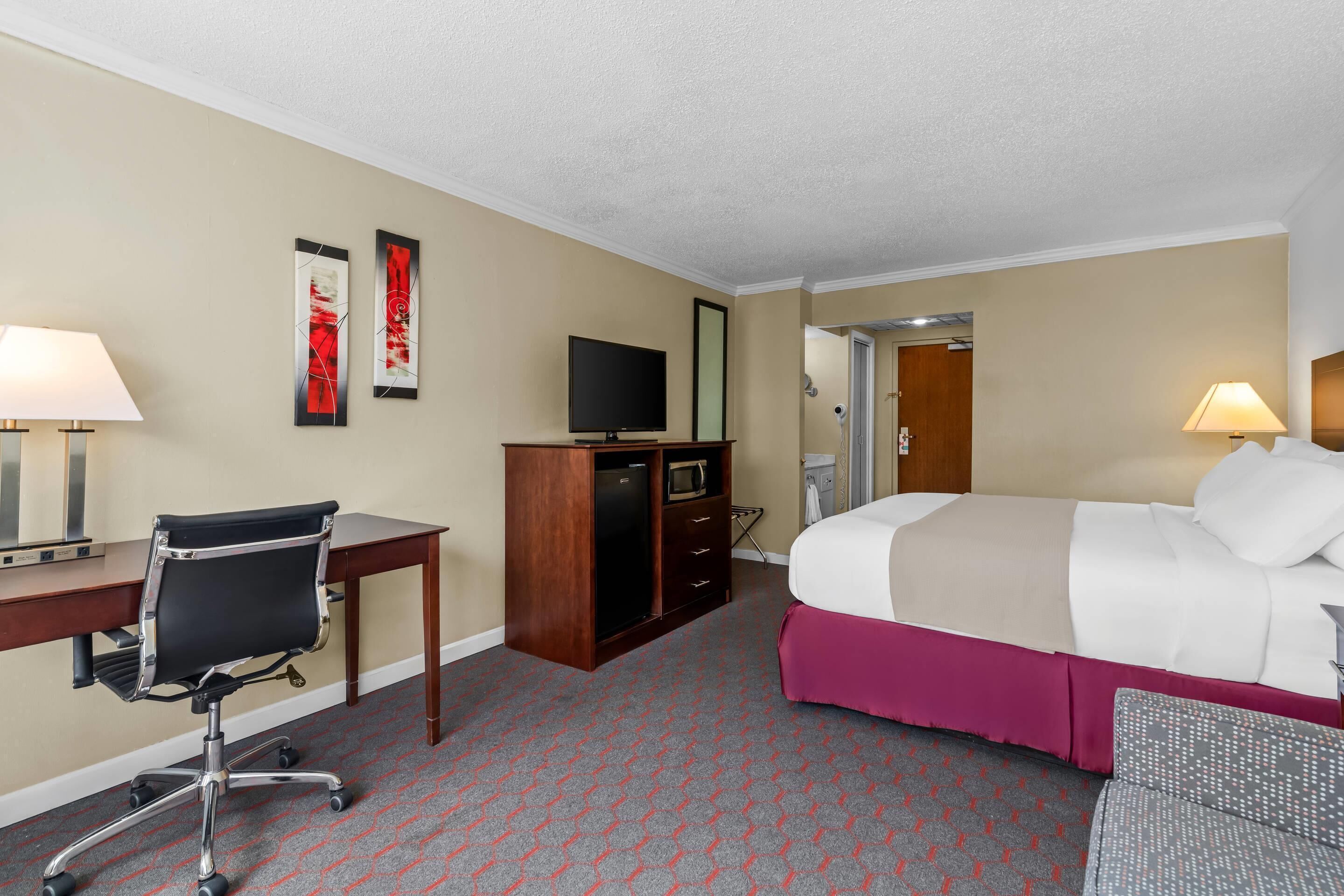 Habitación de hotel con cama, escritorio, silla, televisor, armario y una puerta que da a otra habitación.