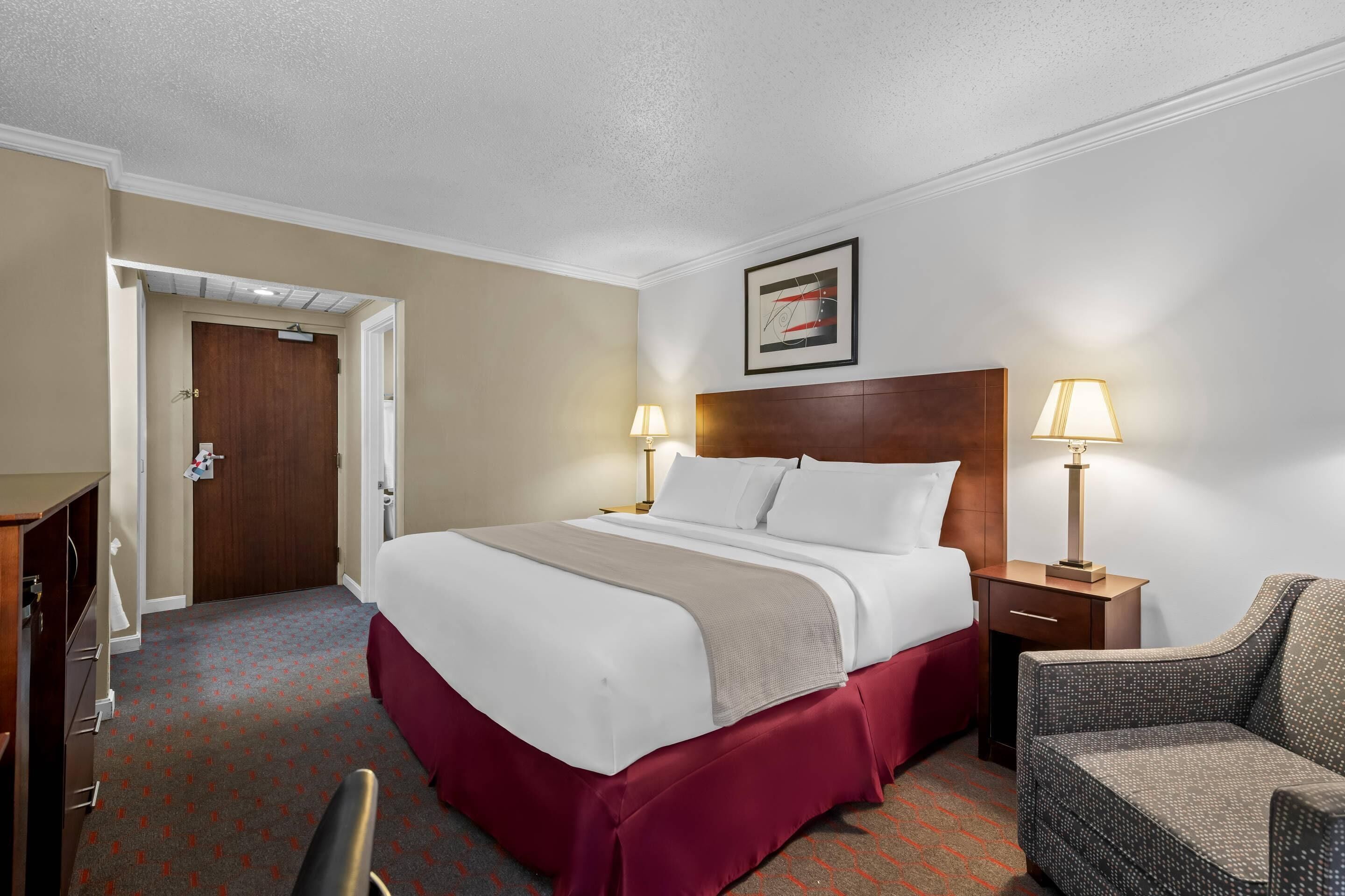 Una habitación de hotel con una cama, dos lámparas de noche, una silla, un escritorio y una puerta.