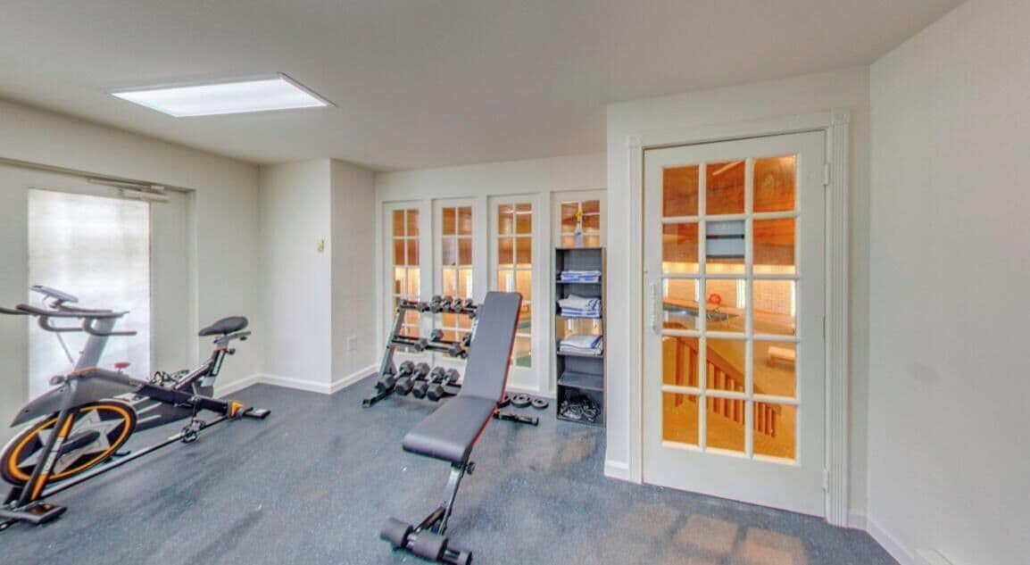 Une salle de sport à domicile équipée de matériel d’exercice, d’un banc, d’haltères et d’un escalier menant à un niveau supérieur.