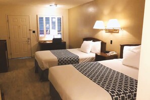 A hotel room with two beds, a desk, a chair, a lamp, and a window with blinds.