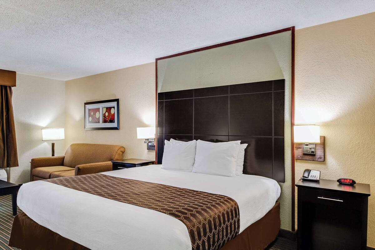 A hotel room with a large bed, a brown armchair, a desk with a lamp, and a nightstand with a phone and clock.