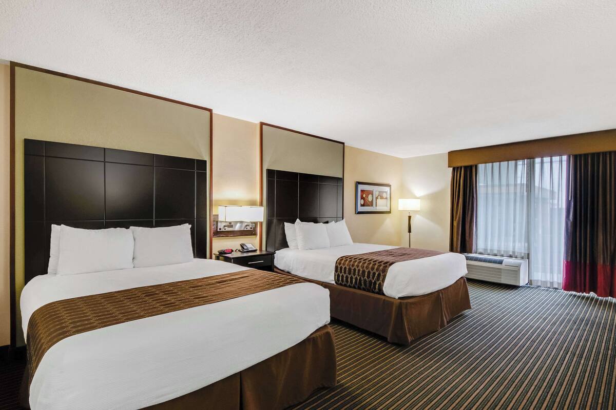 A hotel room with two beds, a large mirror, a nightstand, a lamp, and a window with curtains.