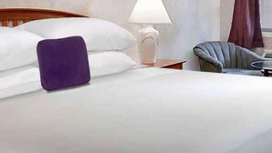 A neatly made bed with white linens and a purple pillow, a wooden headboard, a bedside lamp, and a chair with a round table. - Knights Inn Shreveport (Shreveport)