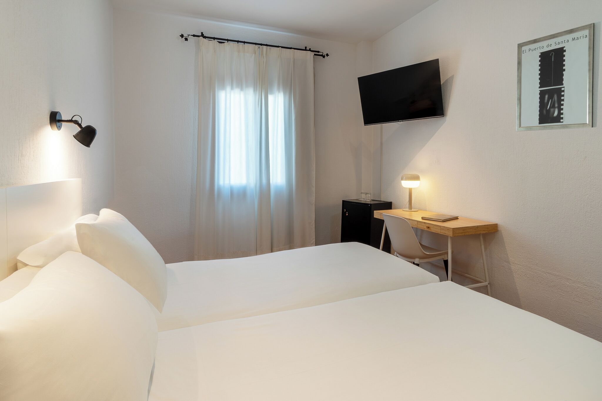 Ein Hotelzimmer mit Bett, Schreibtisch, Fernseher, einem Fenster mit Vorhängen und einem gerahmten Bild an der Wand.