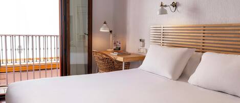 A hotel room with a bed, a desk with a lamp, a chair, and a view of the outside balcony.
