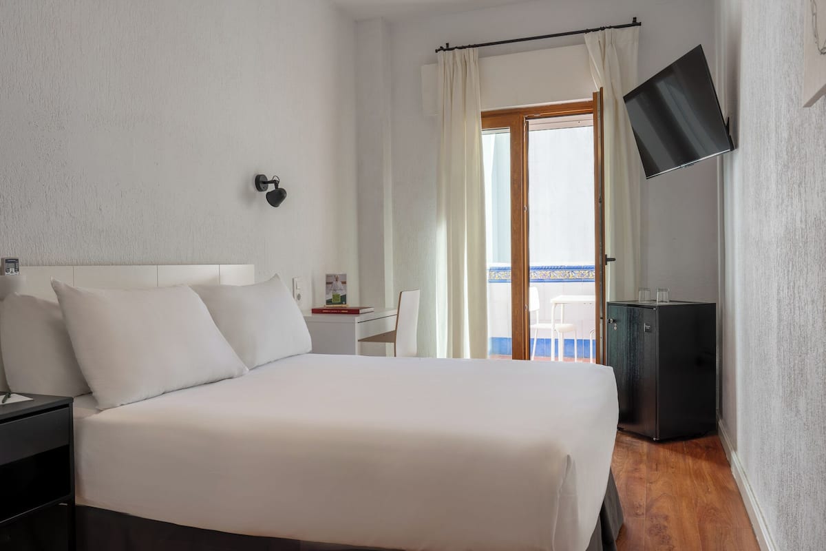 Habitación de hotel con cama, escritorio, silla, televisión y balcón con vistas.
