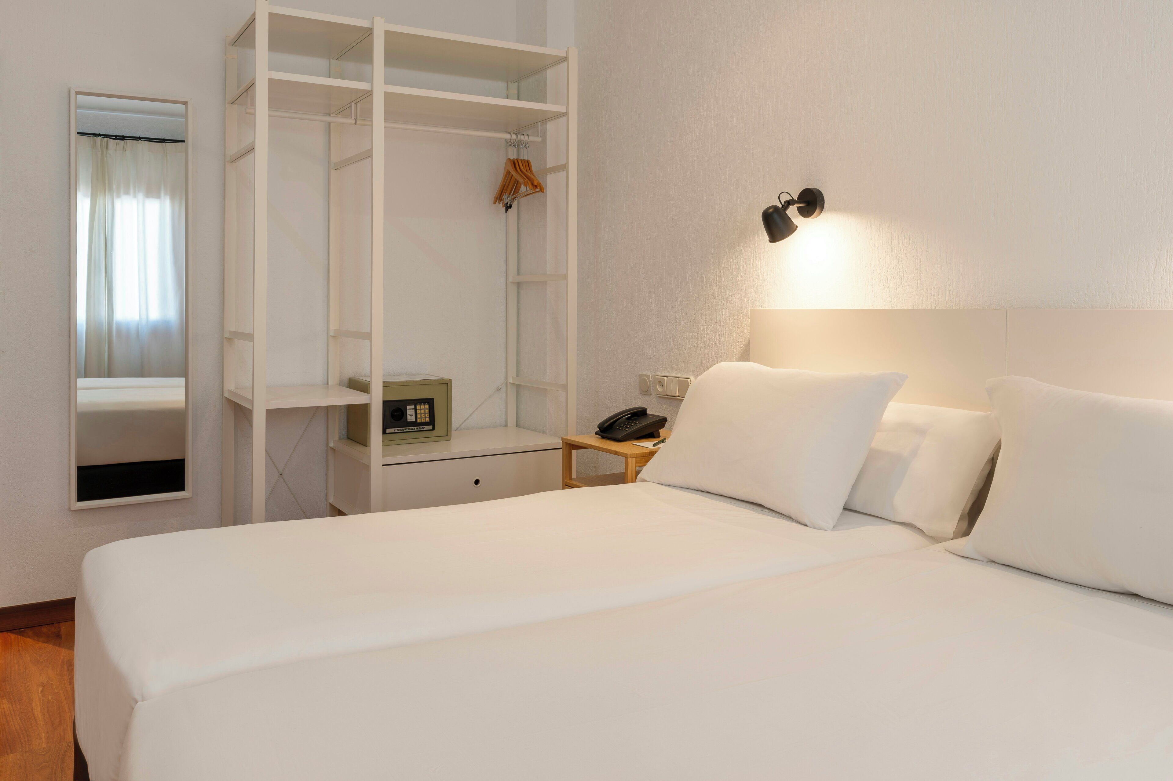 A hotel room with a bed, a bedside table with a phone, a small table with a radio, a wardrobe, and a wall-mounted lamp.