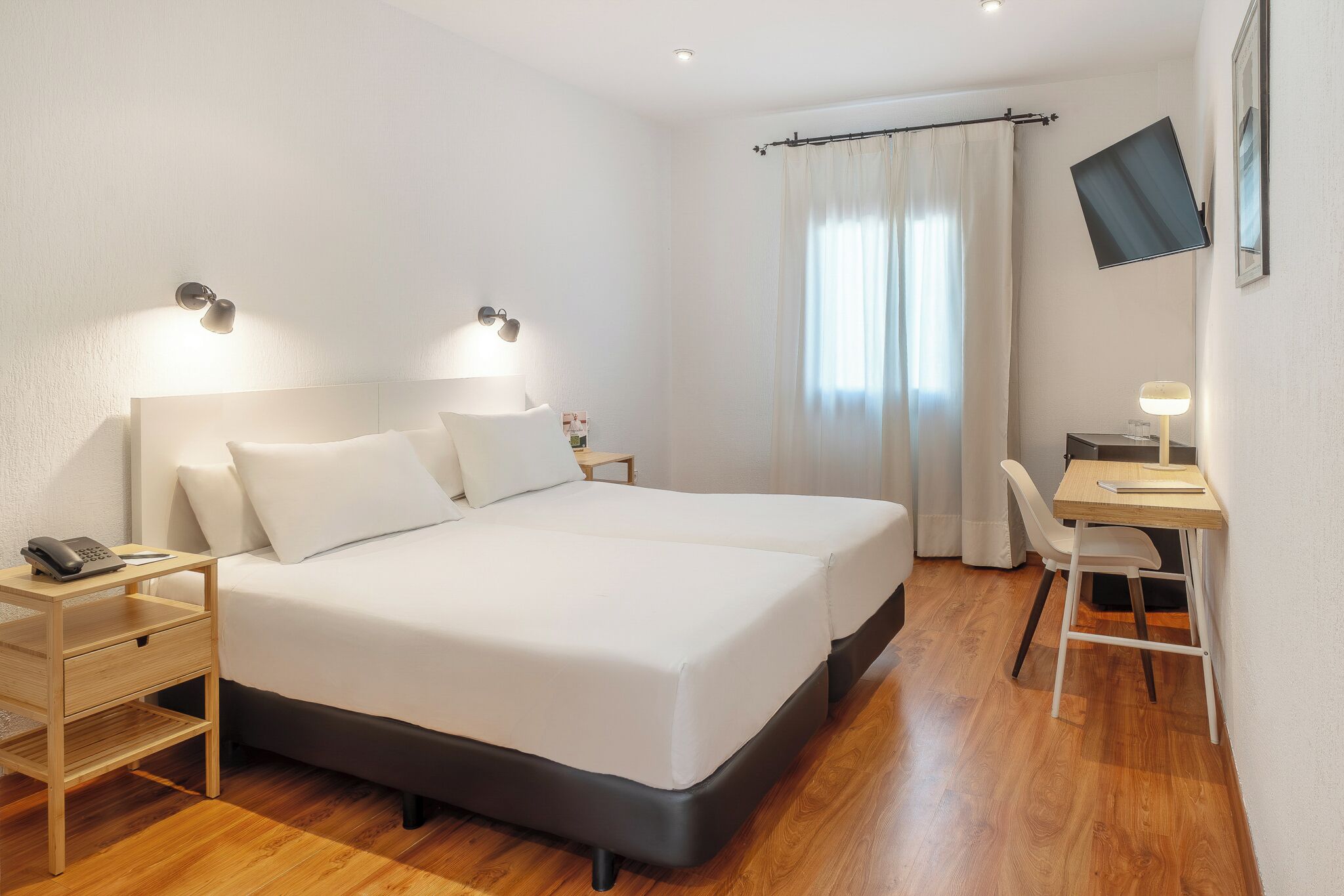 Een hotelkamer met een bed, nachtkastje, bureau, stoel en een aan de muur bevestigde televisie.