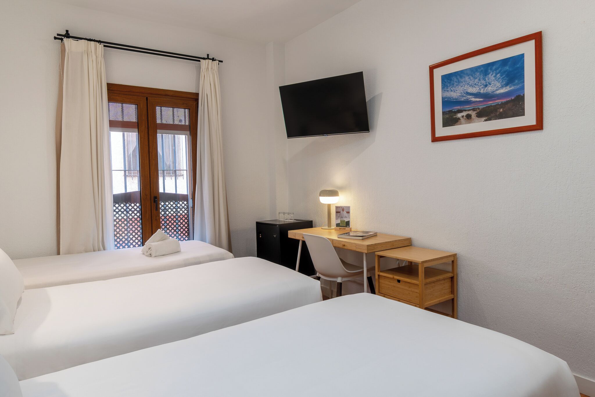 Habitación de hotel con dos camas, un escritorio con silla, televisor montado en la pared y una ventana con cortinas.