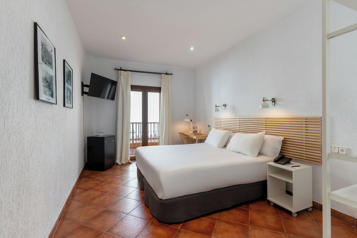 Una habitación de hotel con cama, televisor montado en la pared, escritorio con silla, un mueble pequeño y ventana con cortinas.