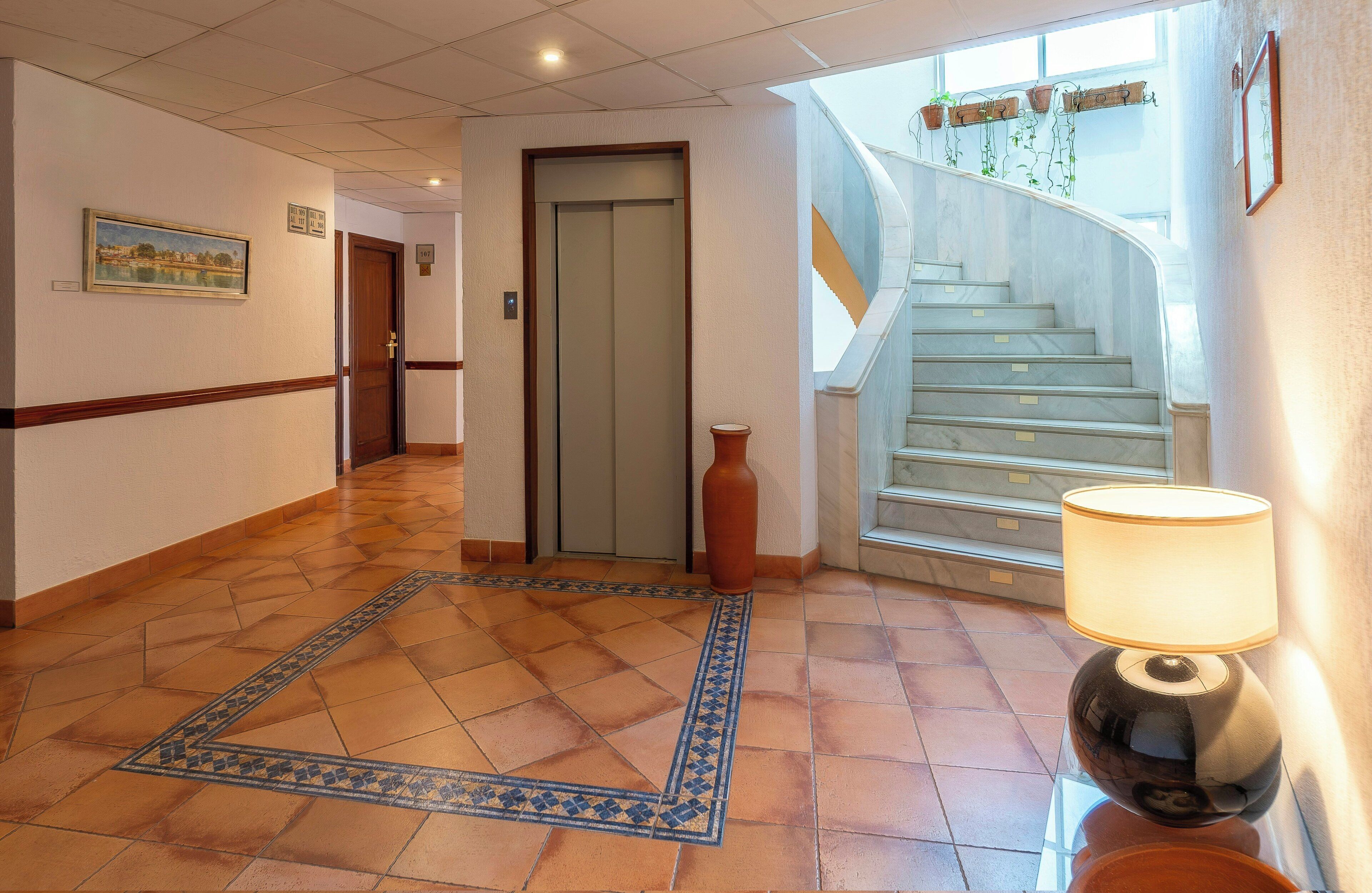 Vestíbulo de hotel con suelo de baldosas, una puerta, una escalera y una lámpara fijada a la pared.
