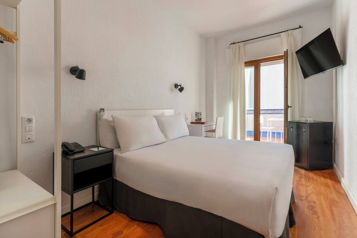 Habitación de hotel con cama, mesita de noche, televisor y balcón con vistas.