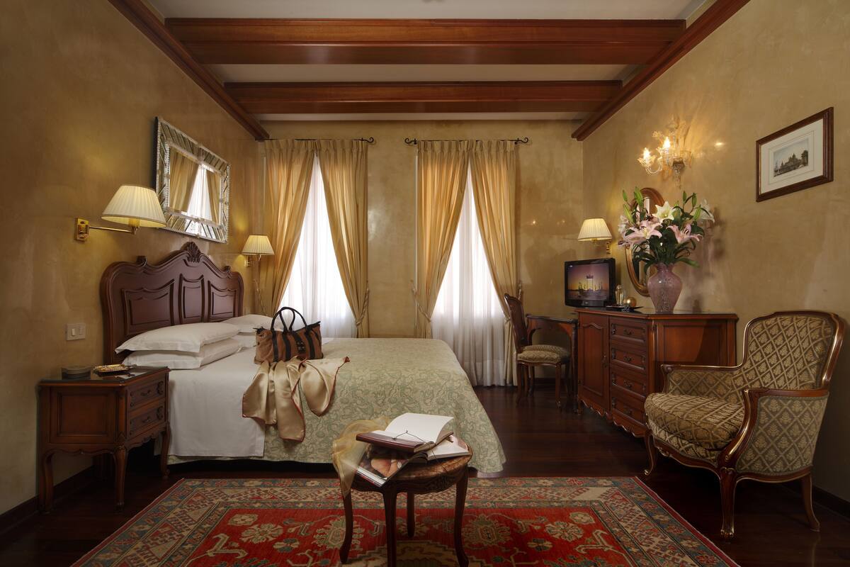 A hotel room with a large bed, wooden headboard, two bedside lamps, a dresser, a chair, a small table with books, and a vase with flowers.