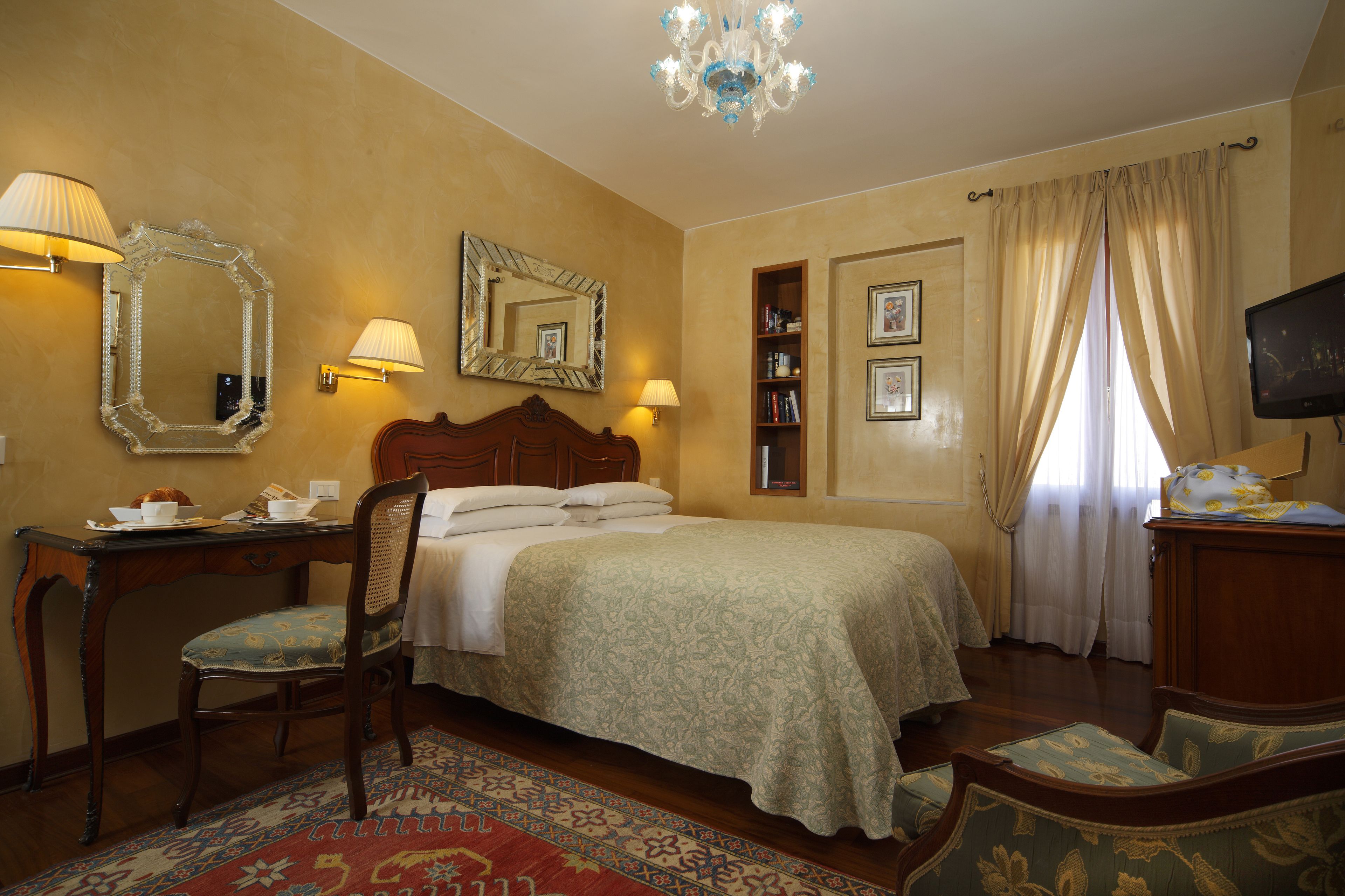 A hotel room with a bed, a desk, a chair, a television, and a bookshelf.