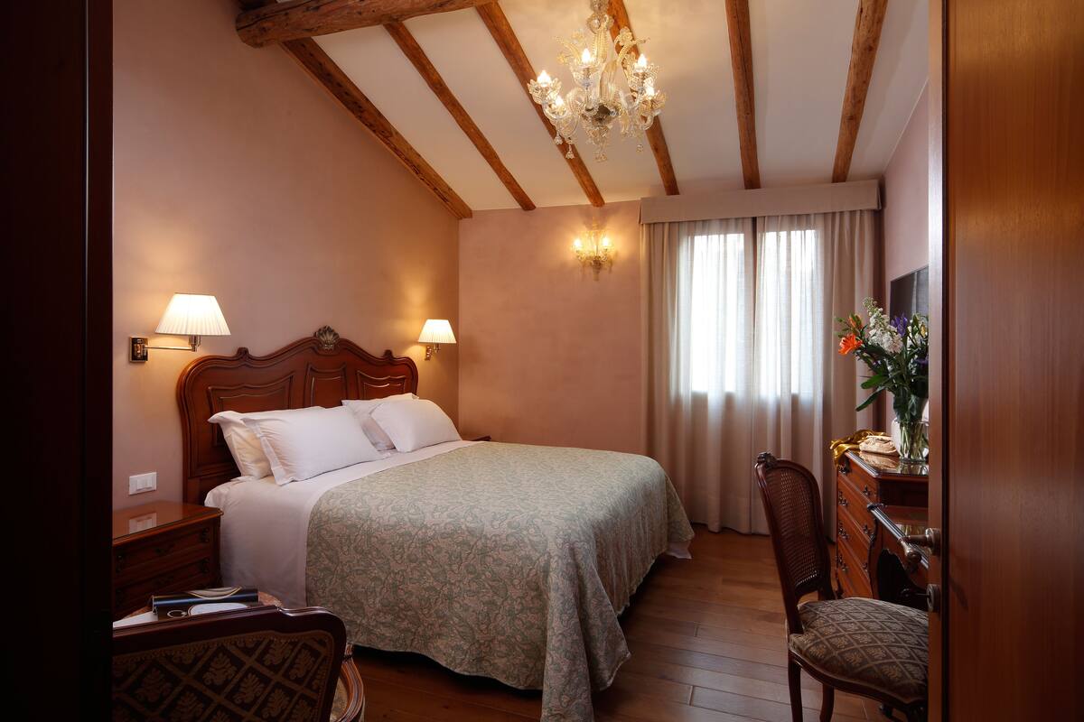 A hotel room with a wooden ceiling, a bed with a patterned bedspread, two bedside lamps, a wooden dresser, and a chair.