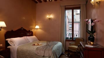 A hotel room with a wooden bed, two bedside lamps, a chair, a desk with a vase, and a window with a view of buildings.