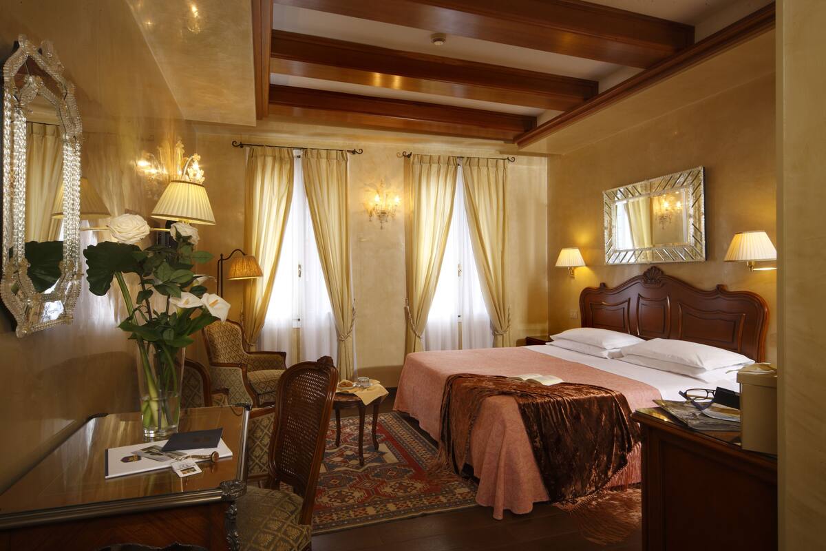 A hotel room with a wooden bed, a desk with a lamp, a chair, a mirror, and a vase with flowers.
