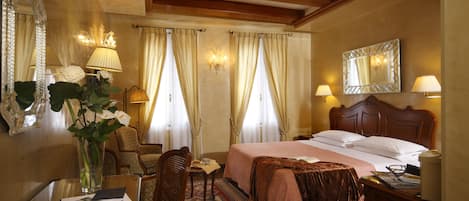 A hotel room with a wooden bed, a desk with a lamp, a chair, a mirror, and a vase with flowers.
