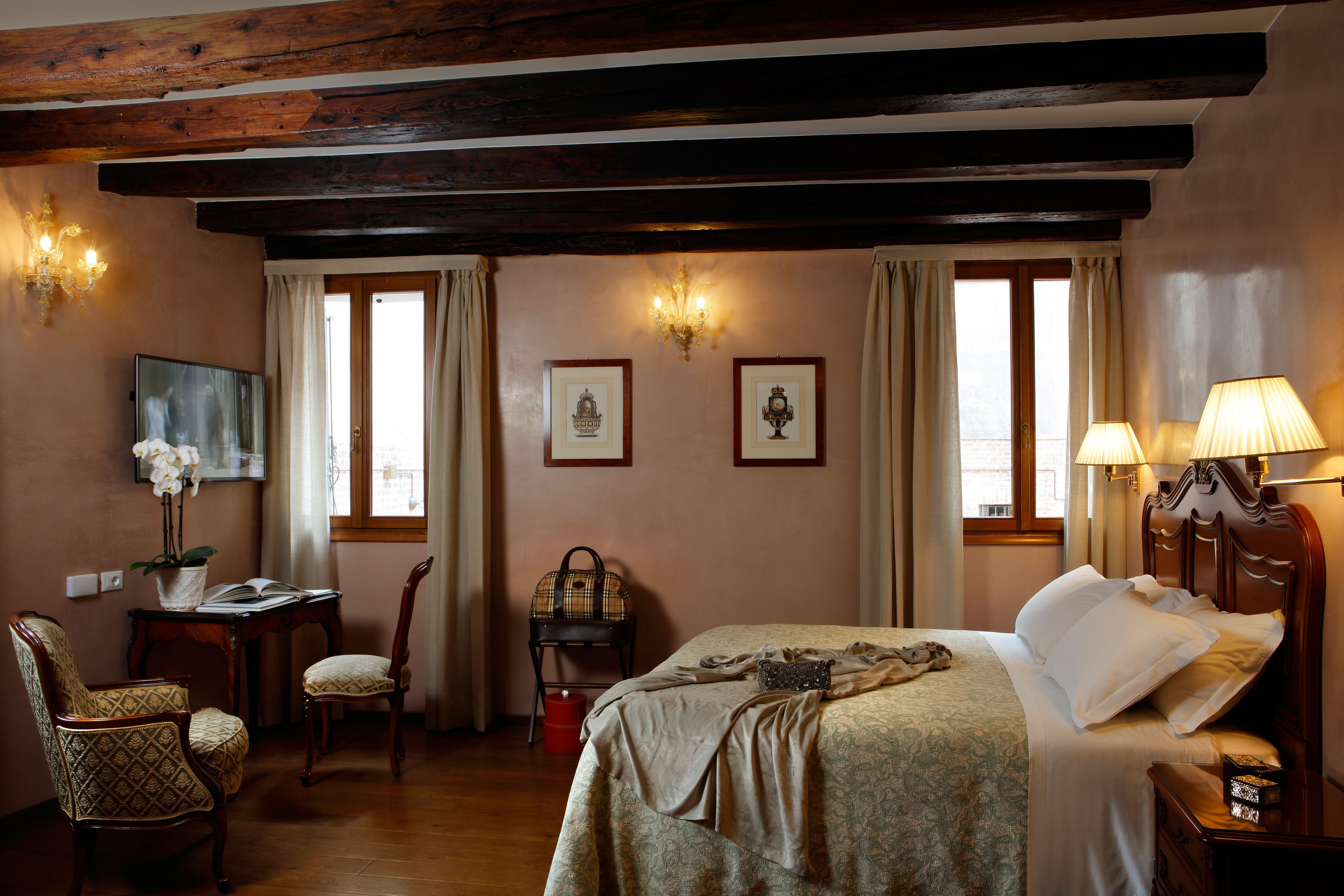 Habitación de hotel con techo de madera, cama con ropa blanca, escritorio con lámpara, una silla y una mesita con un jarrón de flores.